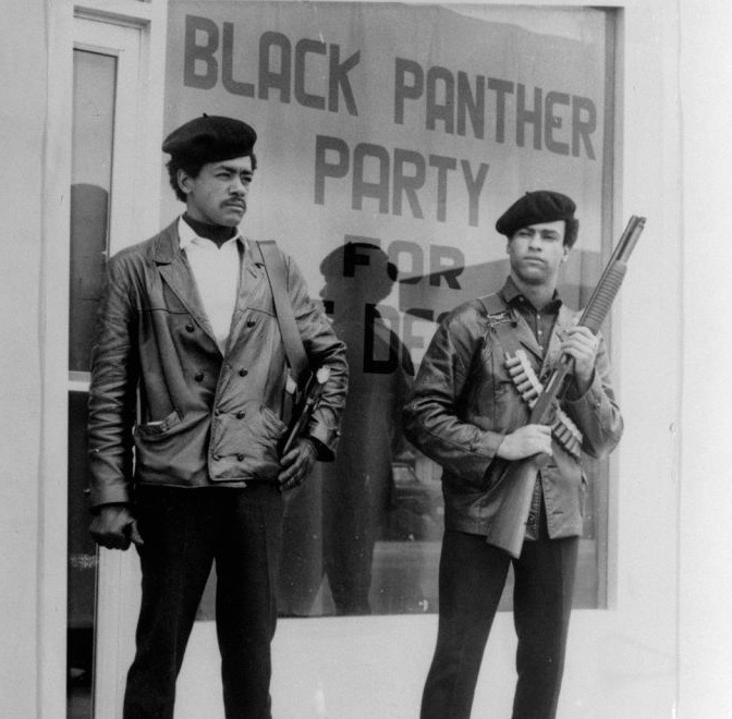 Bobby Seale and Huey Newton standing in front of a sign that reads "Black Panther Party for Self Defense"