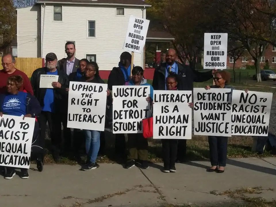 Student and community members protest for the right to literacy, holding signs that have messages like "literacy is a human right."